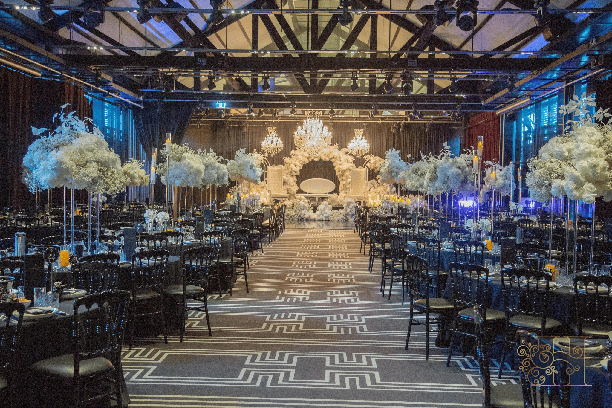 Elegant Indian wedding setup with large white floral centrepieces, a patterned aisle, and ambient blue lighting.